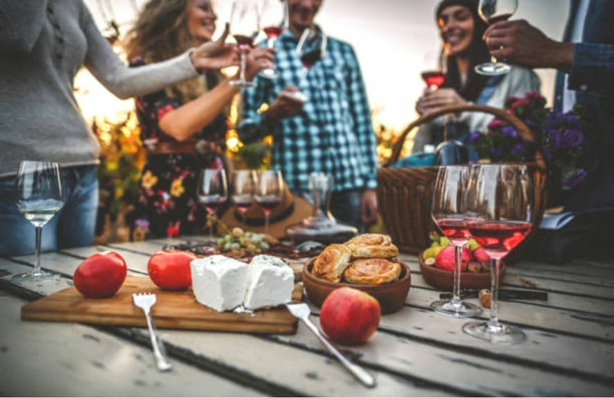 picnic table with glasses of red wine, cheese, fruits and other snacks surrounded by a group of friends