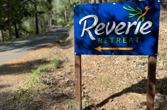 A blue sign reading Reverie Retreat with an arrow pointing left stands beside a shaded forest road surrounded by trees and dry leaves.