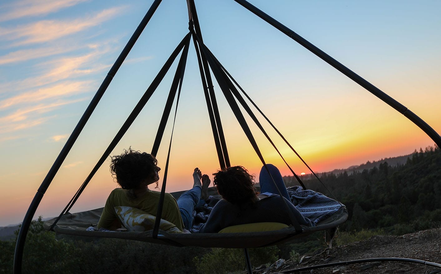 Silhouette of two people relaxing in a large hanging daybed, watching the sunset over a forested valley.