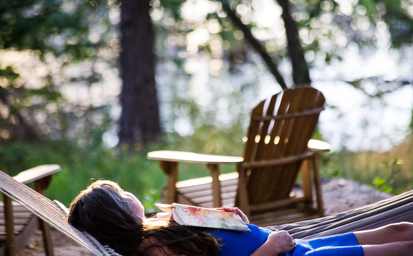 A person relaxing in a hammock with a book, with wooden Adirondack chairs and a sunlit lake in the background.